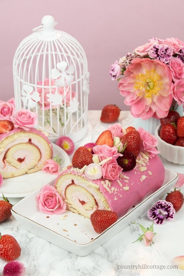 This easy strawberry roll cake recipe features soft sponge cake, a fluffy strawberry cream cheese and whipped cream filling, enrobed by shiny mirror glaze and with fresh berries and flowers as decoration. Whether you call this tasty dessert strawberry cake roll, strawberry Swiss roll, or strawberry roulade, this sweet treat tastes delicious and is perfect for entertaining guests. Strawberry Swiss Roll | Strawberry Roulade #strawberry #rollcake #swissroll #mirrorglaze| countryhillcottage.com