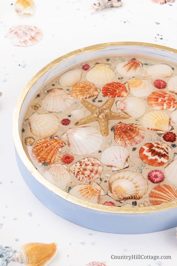 How to make a modern round wood DIY nautical serving tray! The decorative trays idea is a fun summer craft project and rustic farmhouse decor. The step-by-step makeover tutorial includes ideas for materials and working with epoxy resin to create a homemade white painted wooden seashell serving tray with gold trim! Elegant as living room decor, coffee table centerpiece, kitchen display, custom tea tablet, or outdoor party. #servingtray #tray #homedecor #nautical #decor | CountryHillCottage.com