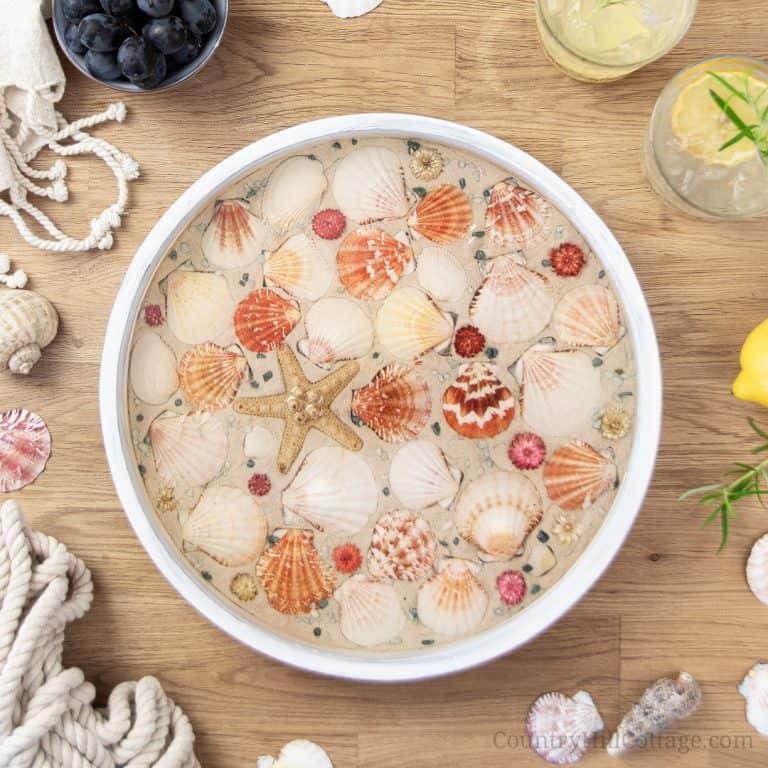 How to make a modern round wood DIY beach tray with seashells! The easy seashell craft is a fun summer craft project and rustic farmhouse coastal decor. The step-by-step makeover tutorial includes ideas for materials and working with epoxy resin to create a homemade white painted wooden seashell serving tray with gold trim! Elegant as living room decor, coffee table centerpiece, kitchen display, custom tea tablet, or outdoor party. #servingtray #tray #homedecor #coastal| CountryHillCottage.com